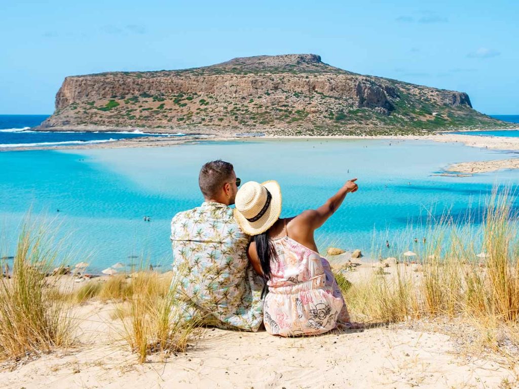 A couple sitting on the sand dunes overlooking Balos Beach in Crete, Greece, enjoying the breathtaking view.