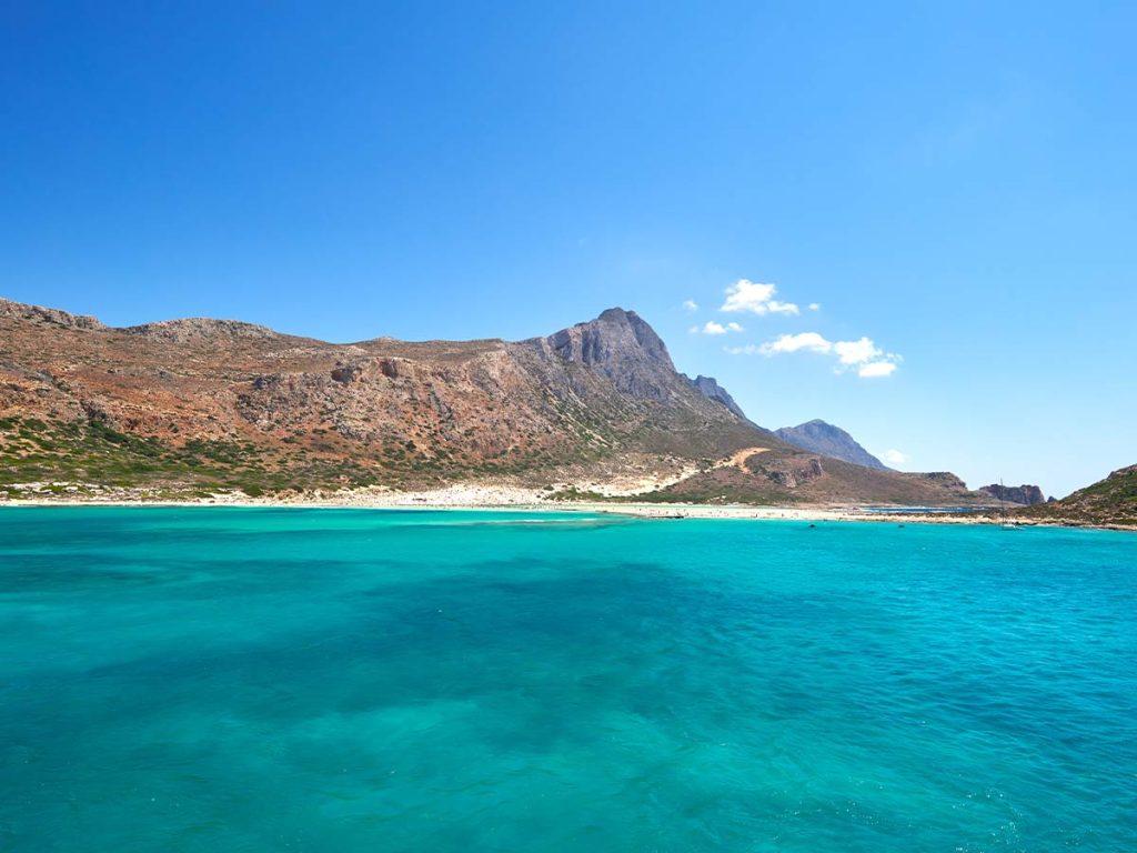 Crystal-clear turquoise waters of Balos Lagoon in Crete, Greece, with mountains in the background under a blue sky.