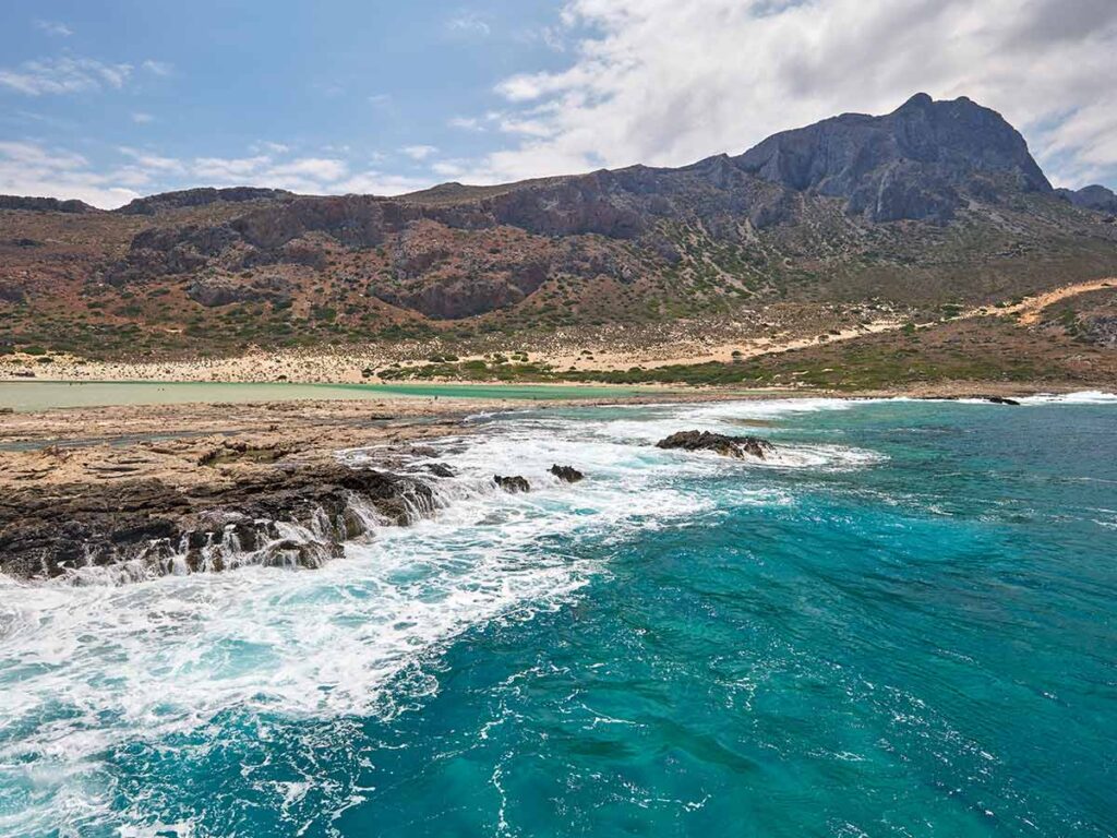 Waves crashing against rocky shore near Balos Lagoon, with mountain backdrop and turquoise sea in Crete.