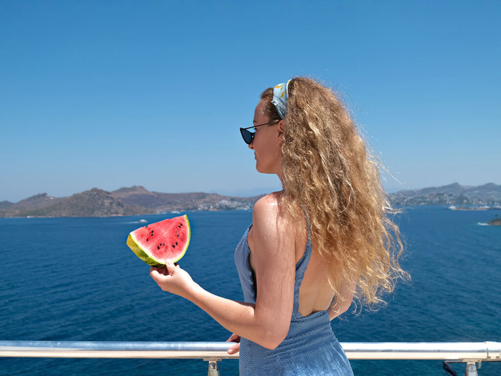 Woman enjoying a slice of watermelon on the deck of a boat with a scenic sea view in Crete – relaxed summer cruise moment.