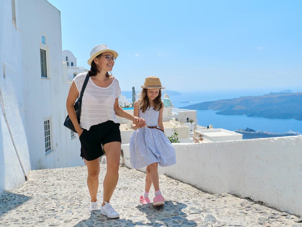Mother and daughter walking on a Cycladic alley with stunning sea view in the background